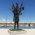 A large sculpture resembling a tree made of metal, with various birds perched on it, set against a waterfront view.