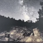 A night sky filled with stars above a rocky landscape with trees in the foreground